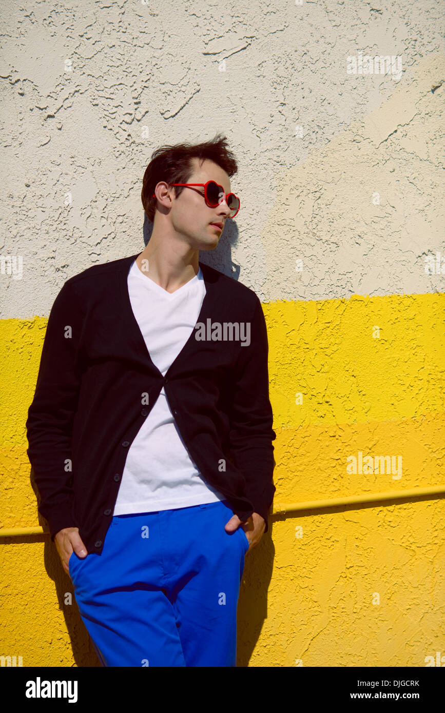 Un giovane uomo, moda modello, vestito in un ambiente informale e blocco di colori moda invernale e il cuore degli occhiali da sole che guarda lontano, appoggiarsi contro la parete. Foto Stock