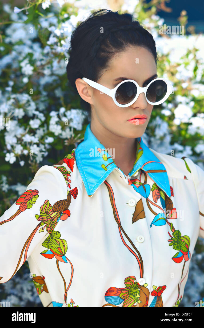 Una bella donna con trecce indossando occhiali da sole rotondi in posa floreali, superiore nella parte anteriore di un fiore bush. Una molla Fashion Concept Foto Stock
