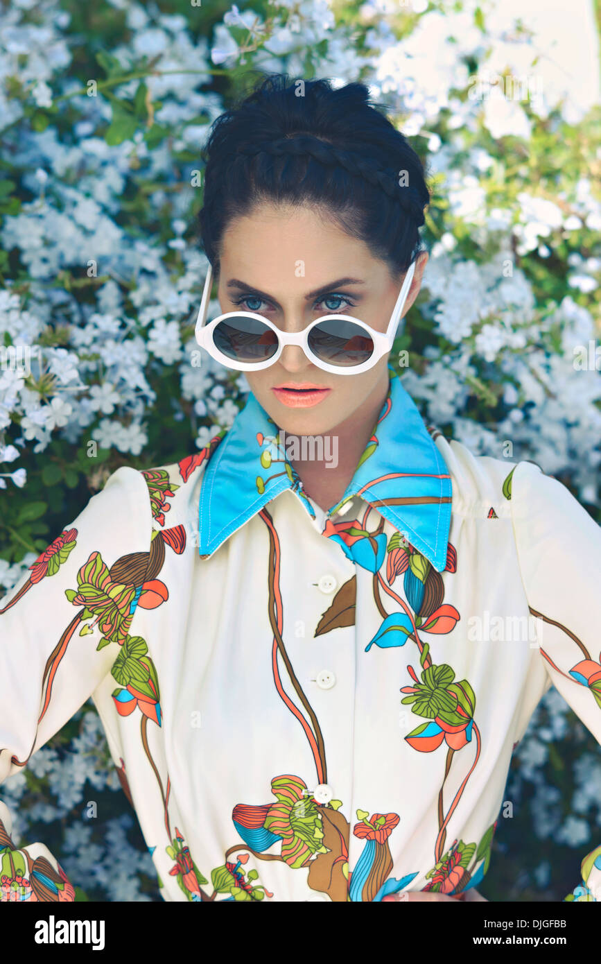 Una bella donna, moda modello in posa davanti a un fiore bush indossando un colorato camicetta floreale e round occhiali da sole. Foto Stock
