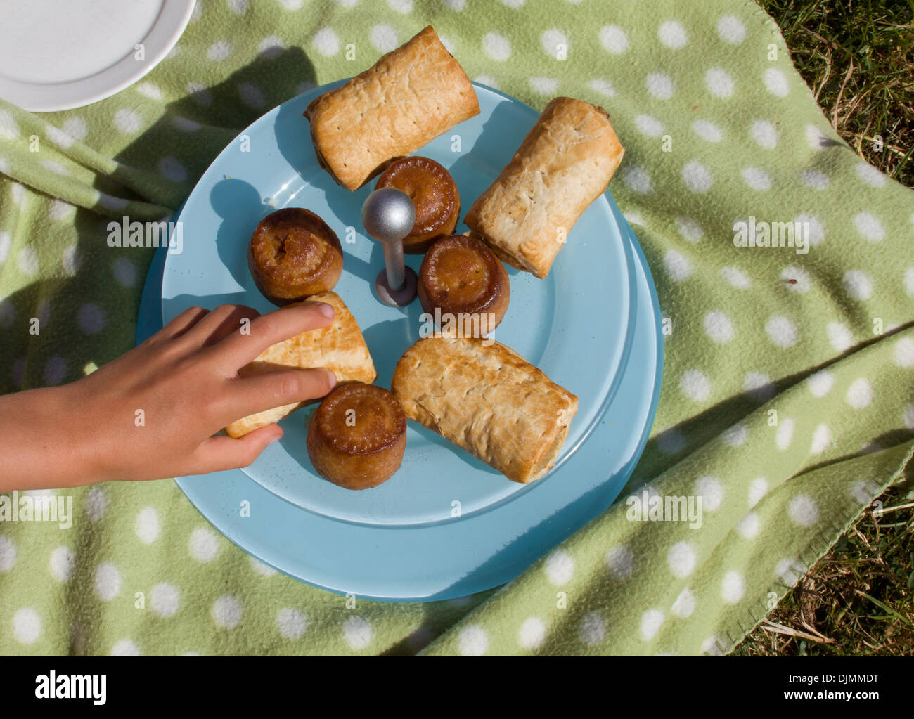 Una chiusura di un bianco caucasico raggiungendo a mano per un rotolo di salsiccia o di carne di maiale torta da una piastra blu su una verde Cesto picnic . Foto Stock