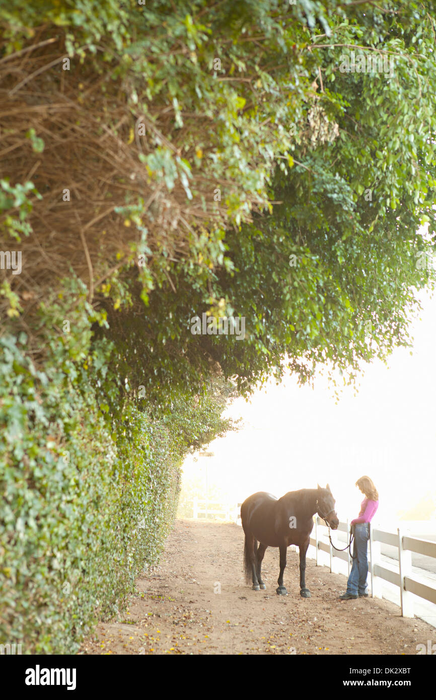 Donna con cavallo marrone appoggiata contro la recinzione di bianco sul percorso di campagna Foto Stock