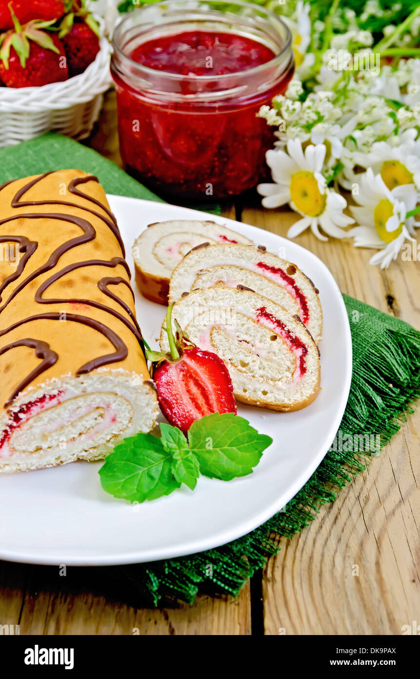 Involtino di biscotto con crema e marmellata, un vasetto di marmellata, tovagliolo, i fiori di camomilla, fragole, menta su una tavola di legno Foto Stock