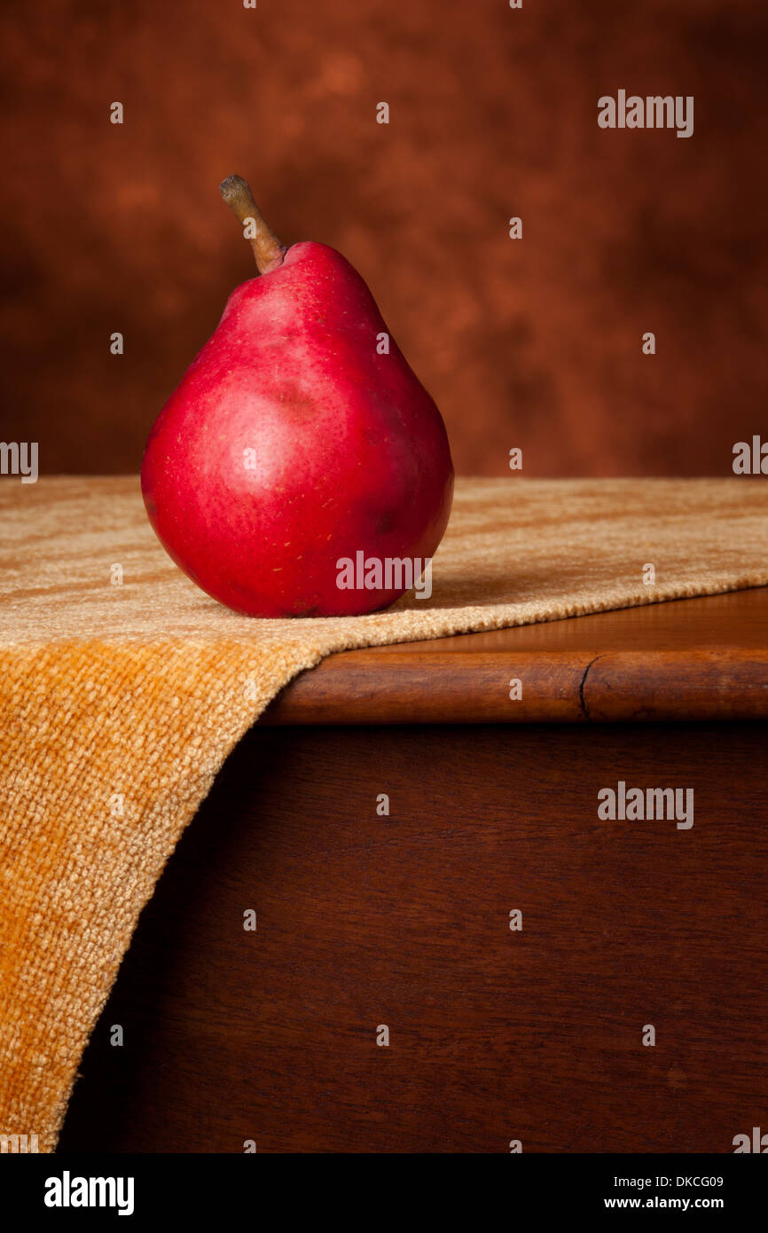 Un singolo red anjou Pera si trova sulla cima di un antico tavolo su un oro tessuto. Questa foto ha calde sfumature di rosso, marrone e oro e Foto Stock