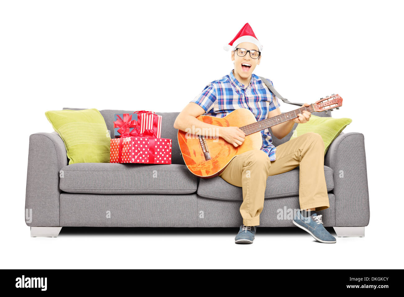 Eccitato giovane maschio con cappello di Natale seduto su un divano moderno e suonando una chitarra acustica Foto Stock