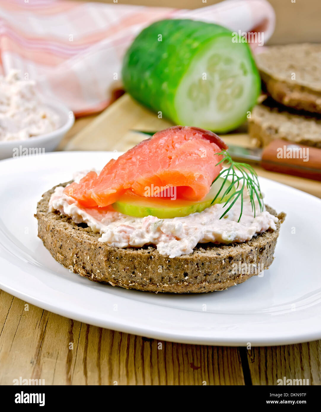 Sandwich di pane di segale con crema, cetriolo, aneto e salmone su una piastra bianca su una tavola di legno Foto Stock