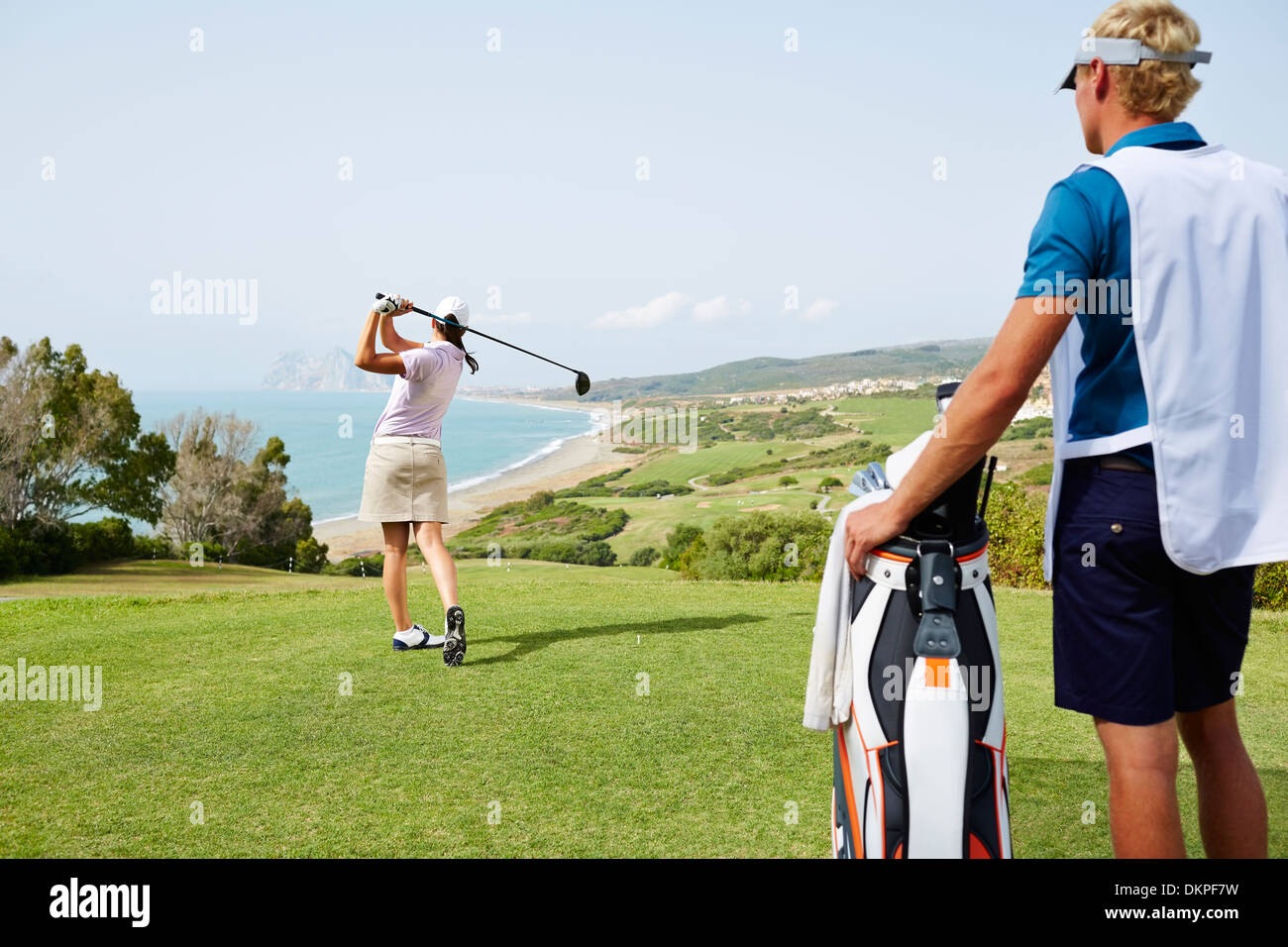 Caddy guardando la donna tee off sul campo da golf con vista sull'oceano Foto Stock