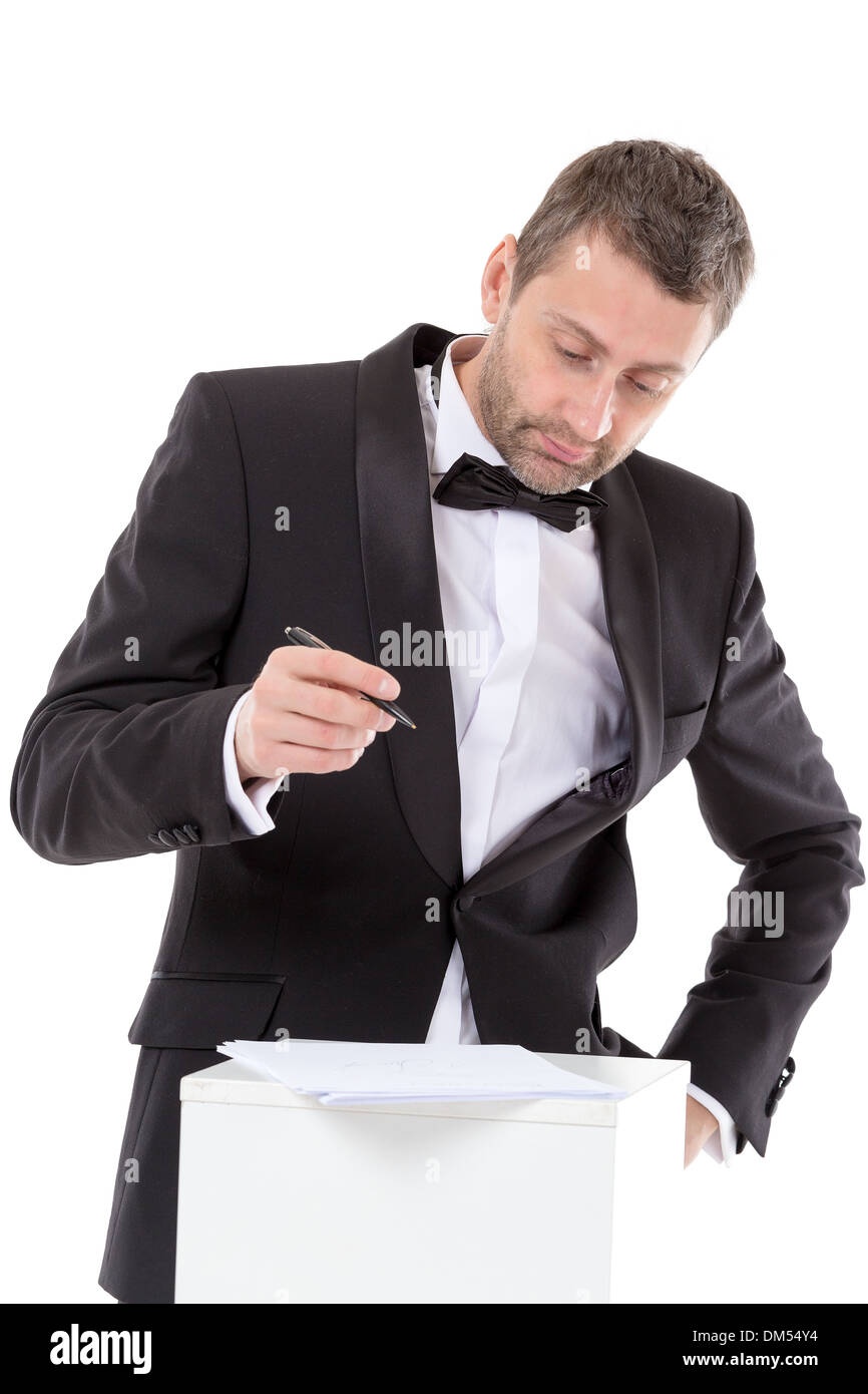 Elegante uomo di mezza età in un bow tie e tuta in piedi in un piedistallo di completare un modulo mettendo in pausa per leggere il documento con una penna Foto Stock