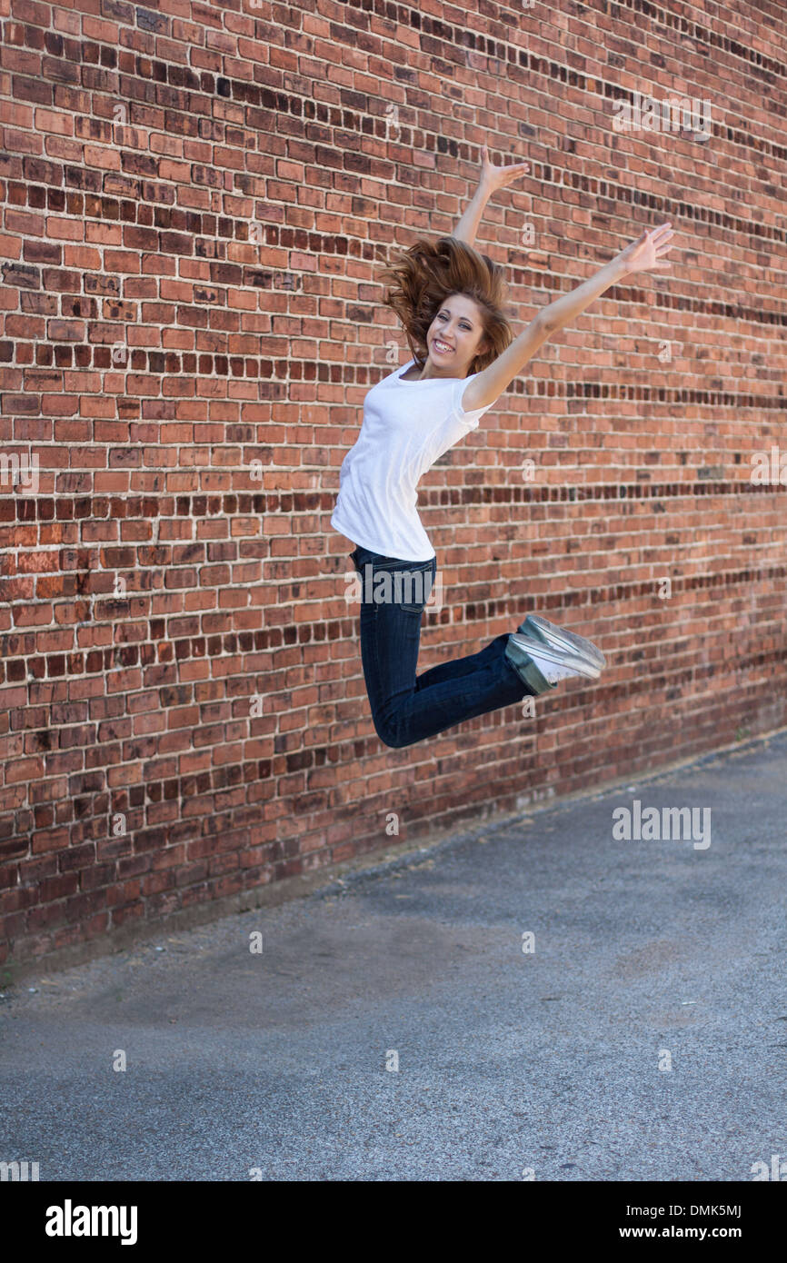 Salti di gioia - orientamento verticale di un felice, sorridente, montare, ragazza adolescente jumping in primo piano, muro di mattoni in background Foto Stock