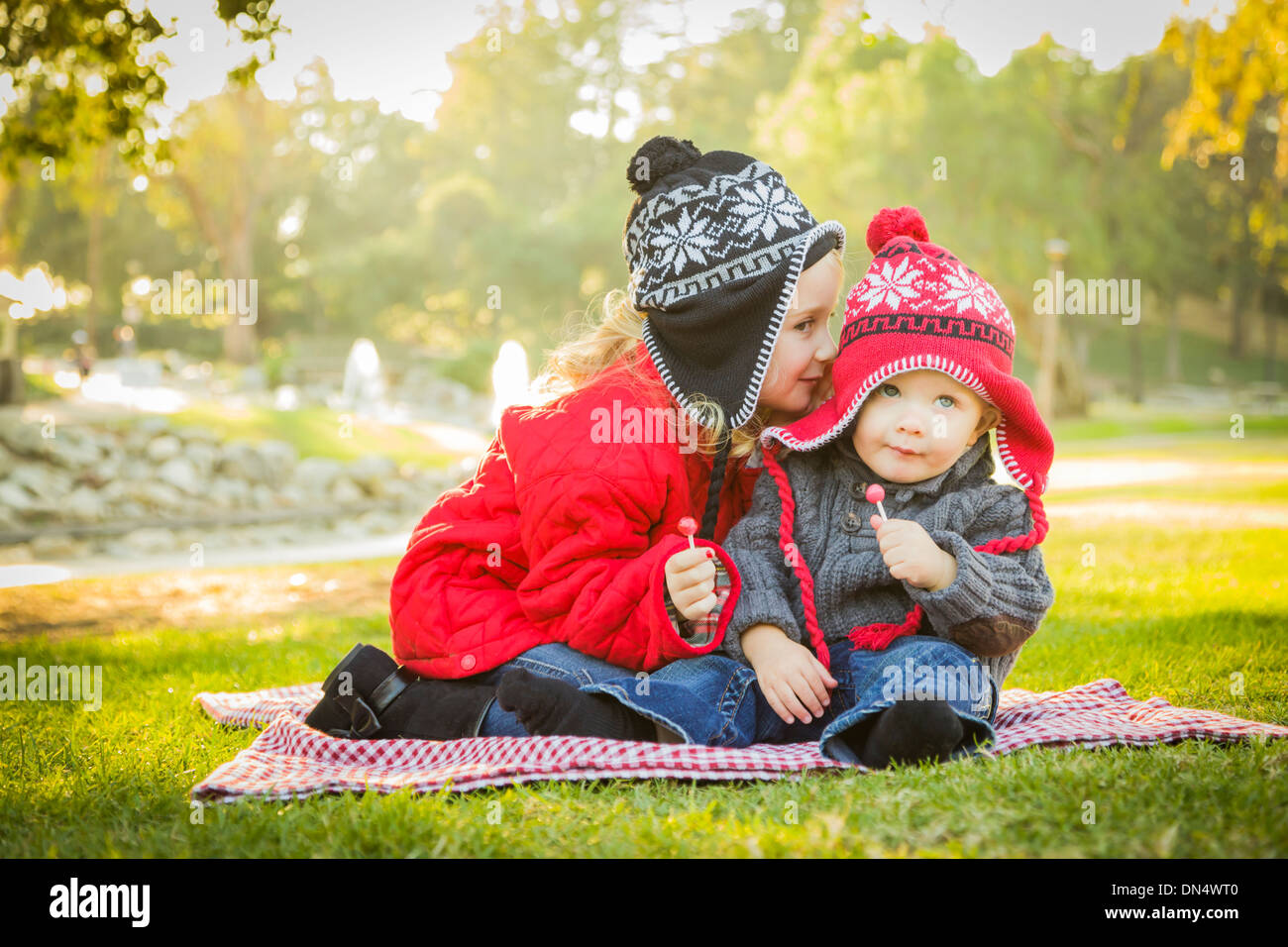 Bambina sussurra un segreto per il suo fratellino indossando cappotti invernali e cappelli seduti all'aperto presso il parco. Foto Stock