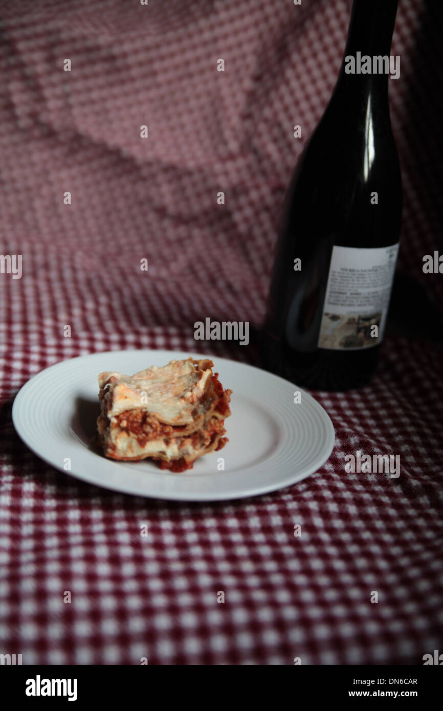 Una fetta di carne fatti in casa lasagne su una piastra bianca con una bottiglia di vino rosso in background in base a una tabella a scacchi panno Foto Stock