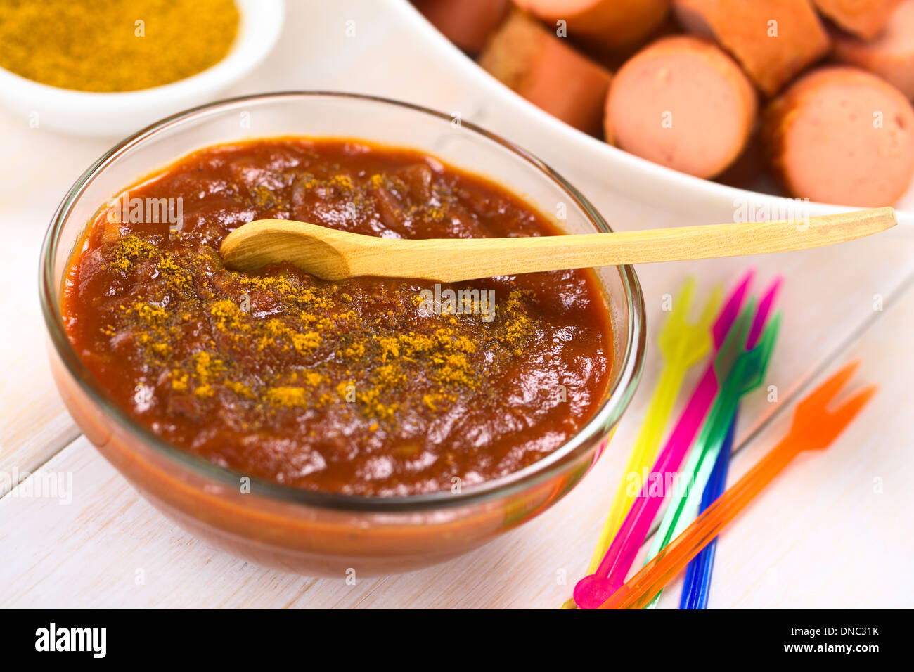 Curry ketchup salsa fatta immergere le salsicce fritte servite in recipiente di vetro con il cucchiaio di legno, plastica colorata parte forche sul lato Foto Stock