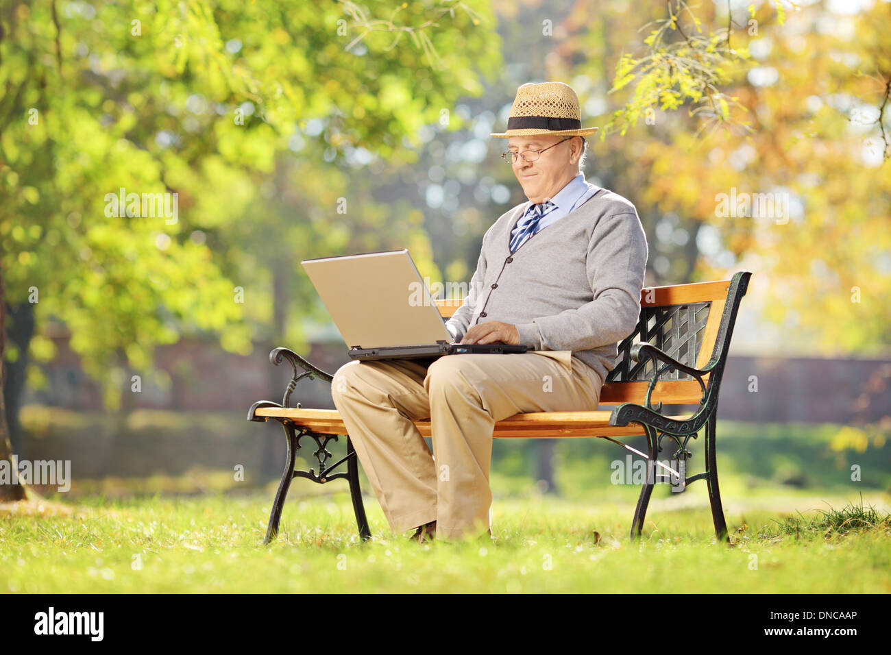 Senior uomo con hat seduta su una panchina e lavora su un computer portatile in un parco Foto Stock