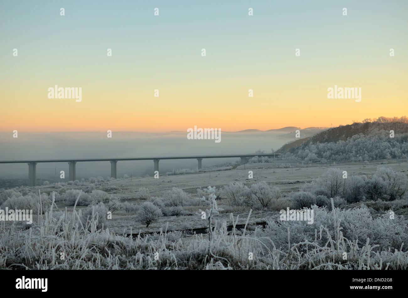 Un inverno alba sopra una fitta nebbia. Sunny alba con cielo blu chiaro oltre il paesaggio innevato. Un ponte può essere visto nella parte anteriore di nebbia. Foto Stock