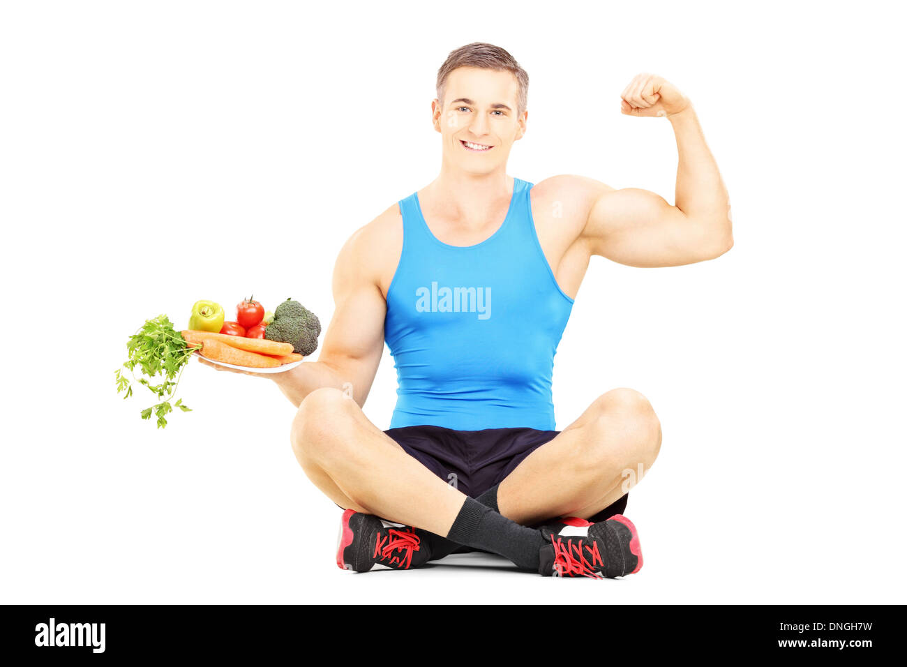 Giovane atleta maschio su un pavimento afferrando un piatto pieno di verdure fresche e mostrando il suo muscolo Foto Stock