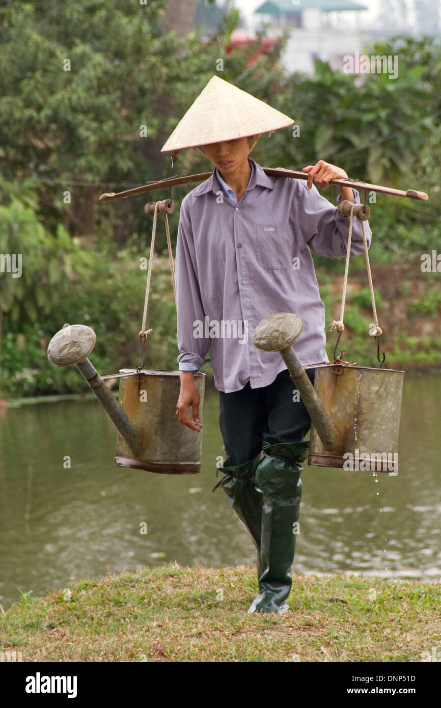 Agricoltore porta grande heavy innaffiatoi riempito di acqua su un palo di spalla dal lago locale di acqua il suo orto. Foto Stock