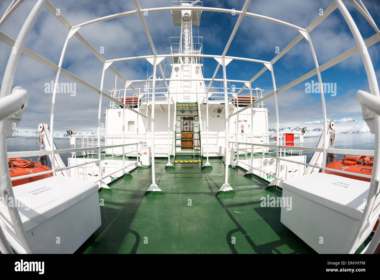 Pontede della nave d'esplorazione Polar Pioneer Vista fisheye Baia Fournier Antartide | BAIA FOURNIER, Antartide — Una vista fisheye spazia sul ponte anteriore della Polar Pioneer, una nave di Aurora Expeditions, con montagne innevate della penisola antartica che costeggiano l'orizzonte. Le casse sul ponte marcate "CEMENT" e "ПЕСОК" (russo per "sabbia") tradiscono le origini di ricerca dell'era sovietica della nave, costruita in Finlandia nel 1982 prima della conversione per crociere d'esplorazione. Foto Stock