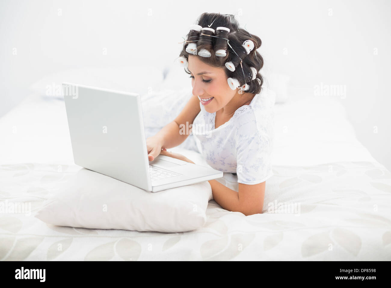 Bella bruna nei capelli rulli sdraiato sul suo letto utilizzando il suo laptop Foto Stock