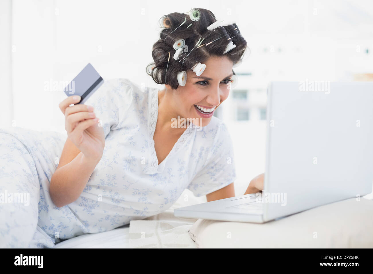 Emozionato brunette nei capelli rulli sdraiato sul suo letto utilizzando il suo laptop per shop online Foto Stock