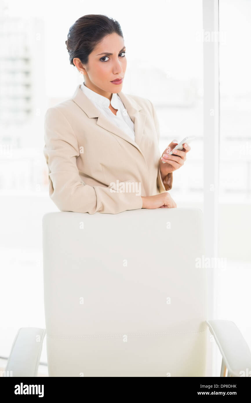 Accigliata imprenditrice in piedi dietro la sua sedia tenendo il suo telefono lampante in telecamera Foto Stock