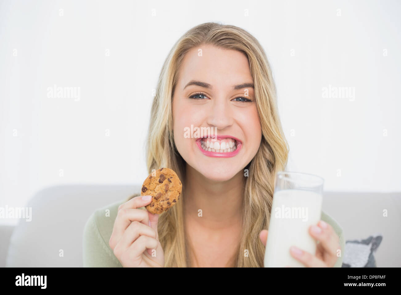 Felice biondo carino mangiare cookie con latte seduto sul divano accogliente Foto Stock