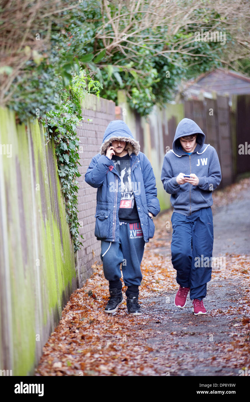 Due ragazzi adolescenti in hoodies camminare insieme verso il basso un vicoletto suburbana texting e parlando con i loro cellulari. Foto Stock