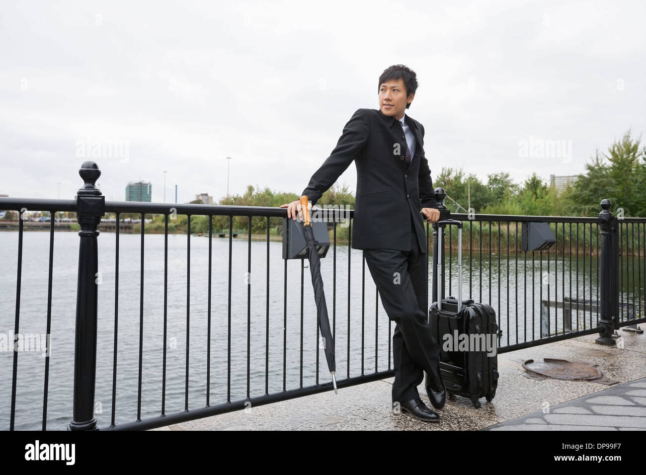 Lunghezza completa di imprenditore con bagagli appoggiata sulla ringhiera lungo il fiume Foto Stock