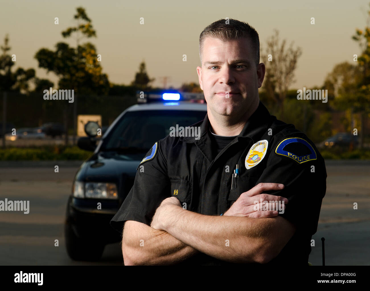 Una forte ricerca funzionario di polizia con le braccia piegate e la sua auto di pattuglia in background. Foto Stock