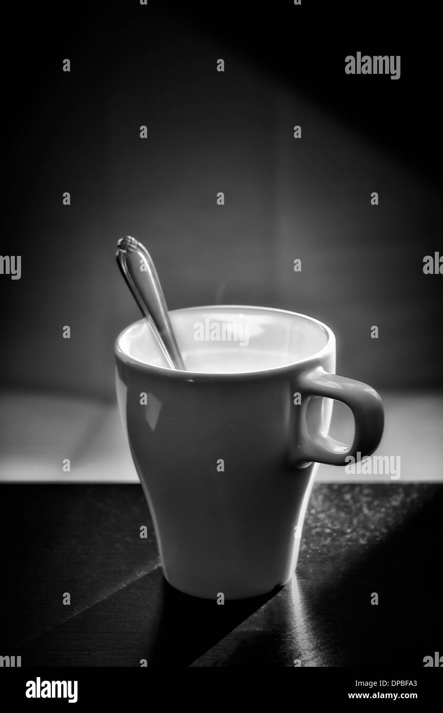 Un bianco tazza di caffè con il cucchiaio su una tavola nera. retroilluminazione luce del sole di mattina Foto Stock