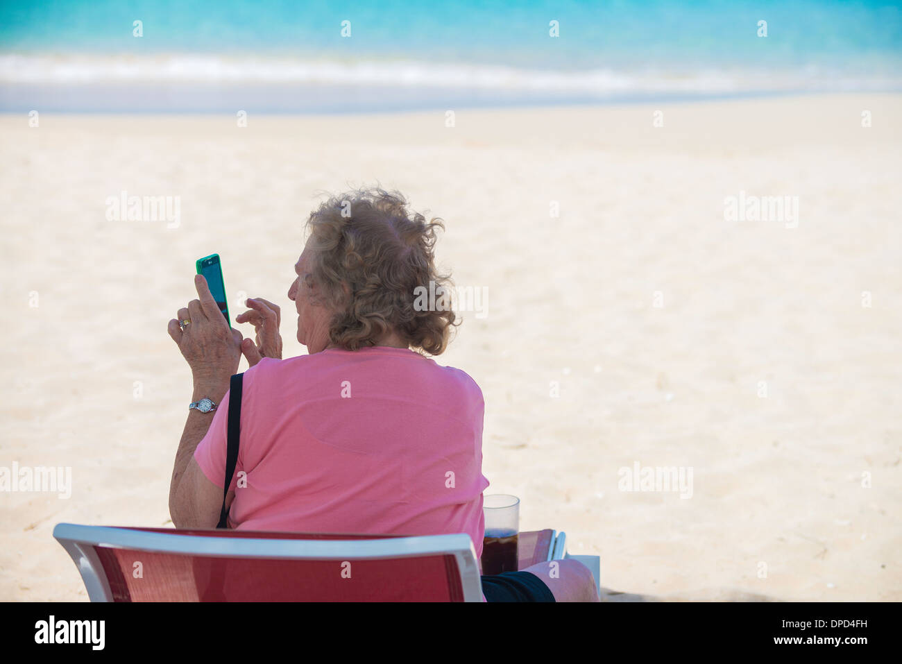 Un'anziana donna caucasica si siede in una sdraio sulla spiaggia sulla spiaggia di Sandcastle, affacciato sul Mar dei Caraibi e gioca con il suo smart phone. Foto Stock Un'anziana donna caucasica si siede in una sdraio sulla spiaggia sulla spiaggia di Sandcastle, affacciato sul Mar dei Caraibi e gioca con il suo smart phone. Foto Stock