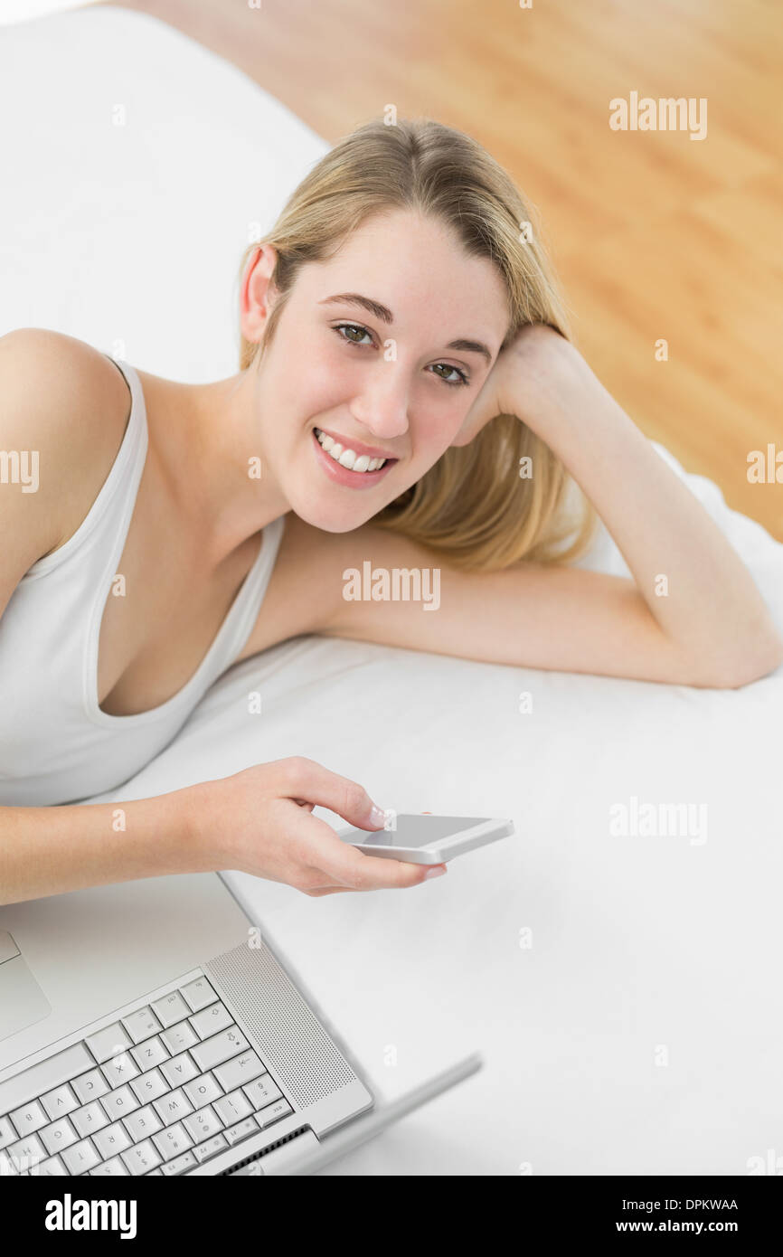 Gleeful donna bionda giacente sul suo letto tramite il suo smartphone Foto Stock