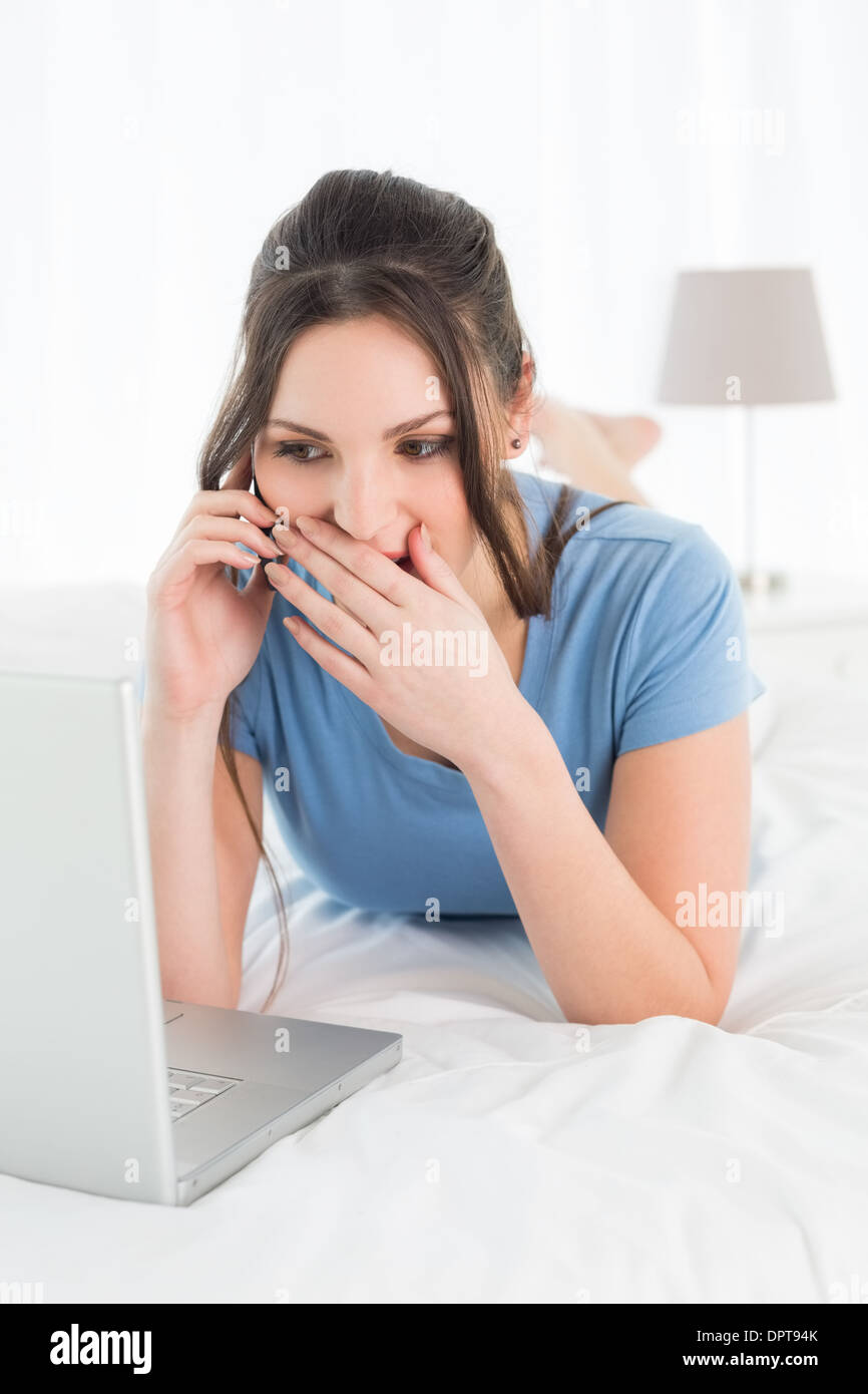 Scioccato donna mediante telefono cellulare e computer portatile a letto Foto Stock