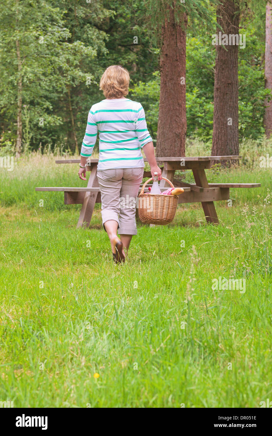 Una donna che porta un picnic di vimini cesto pieno di cibo e bevanda verso una tabella nel bosco su una luminosa giornata d'estate. Foto Stock