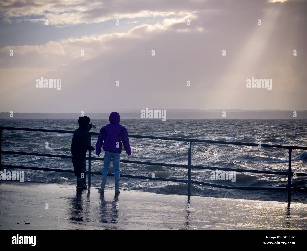 Due bambini di guardare oltre il mare in tempesta come il sole fa capolino tra le nuvole Foto Stock
