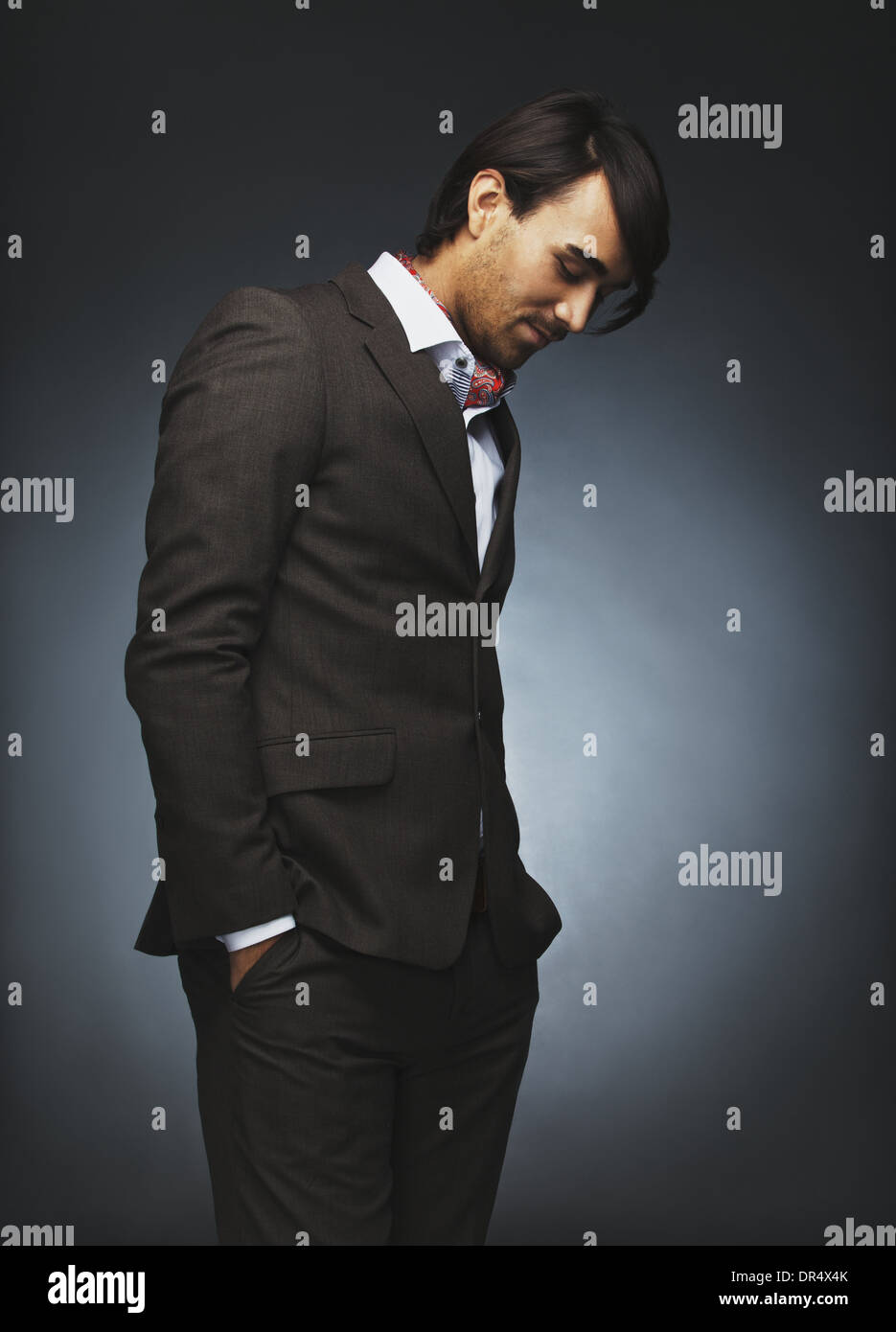 Attraente giovane uomo vestito elegante in piedi su sfondo nero. Moda maschile modello con le mani in tasca guardando verso il basso Foto Stock