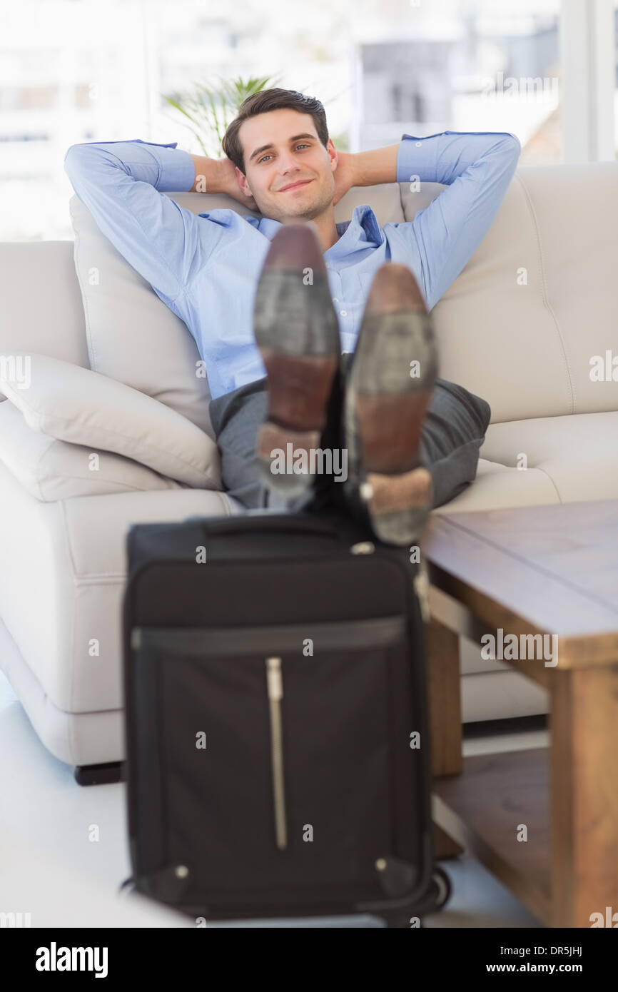 Imprenditore sorridente seduto sul divano con piedi sulla valigia Foto Stock