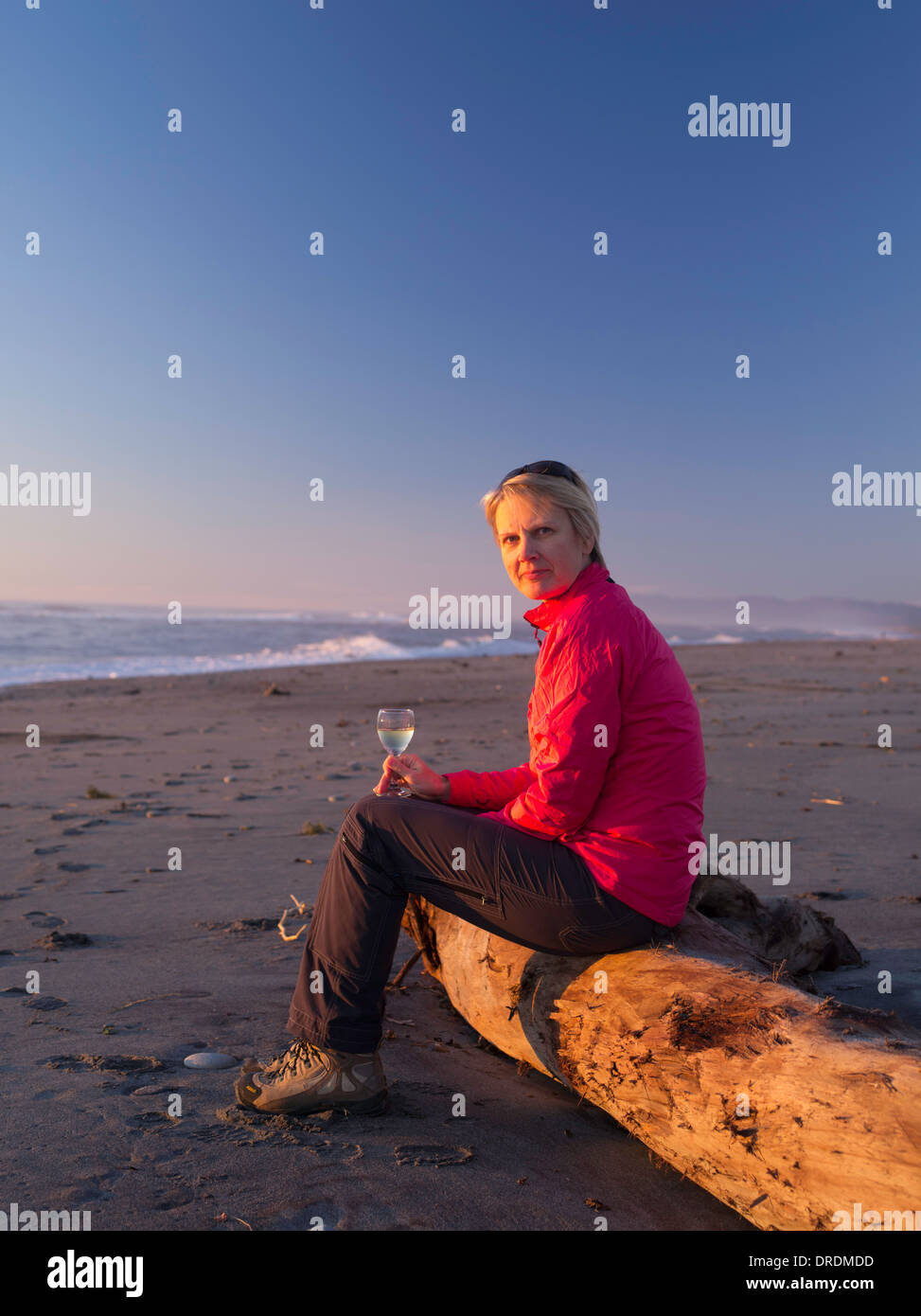 Una di mezza età, donna bionda gode il suo bicchiere di vino bianco mentre è seduto su un registro di deriva in spiaggia, Hokitika, Nuova Zelanda. Foto Stock