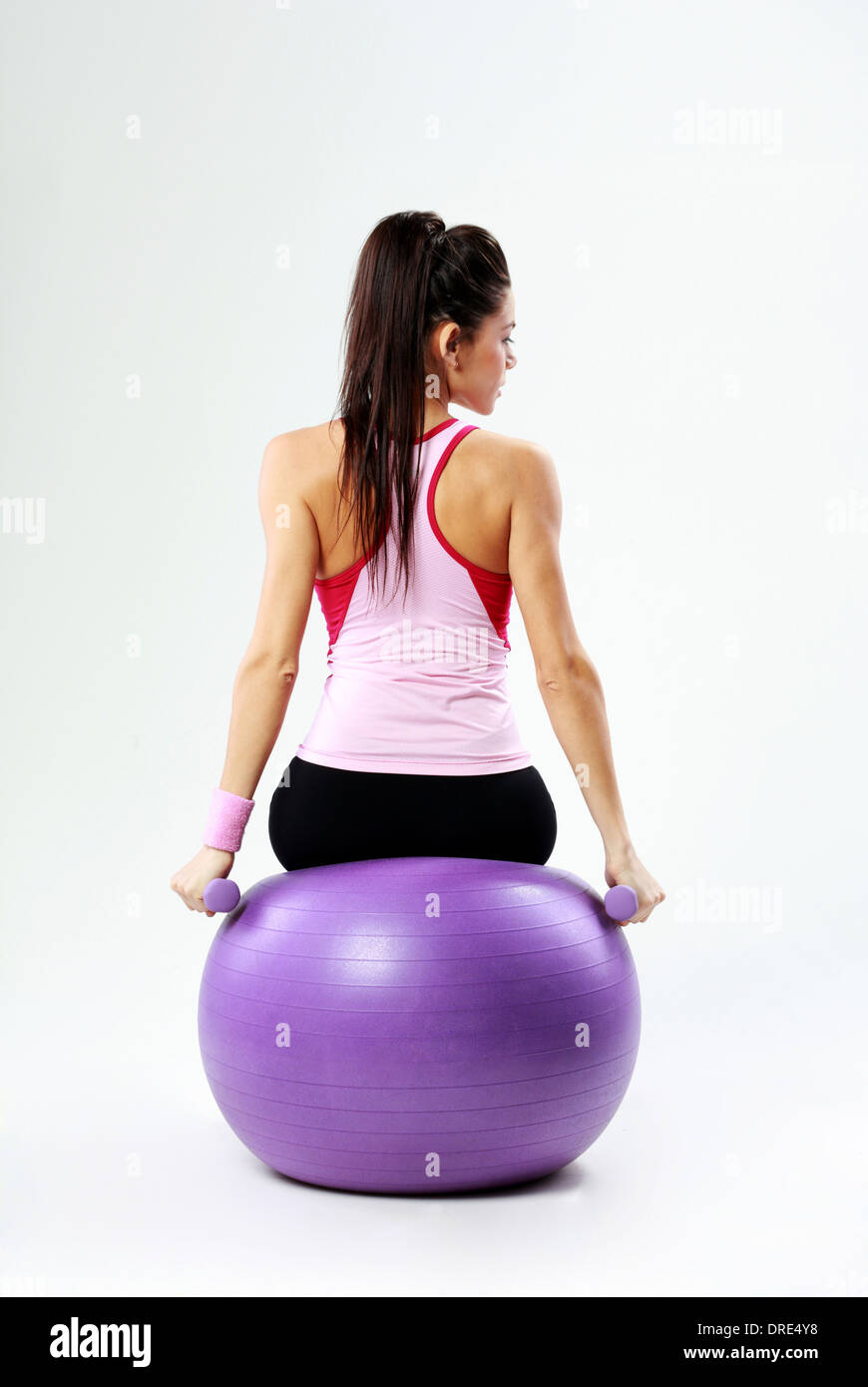 Vista posteriore di una giovane sport donna seduta su fitball con manubri su sfondo grigio Foto Stock