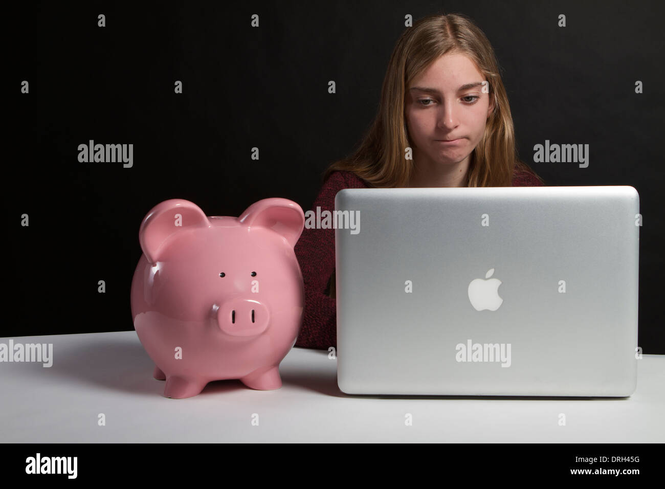 Preoccupato teenage studente di college alla ricerca di un prestito per studenti su un computer con una rosa salvadanaio sulla scrivania accanto a lei . Foto Stock