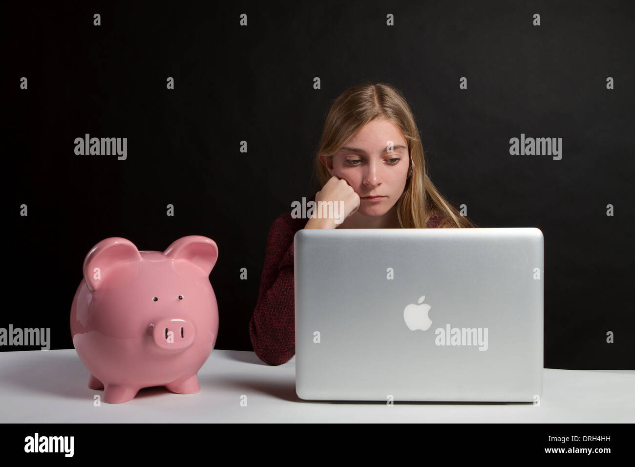 Preoccupato teenage studente di college alla ricerca di un prestito per studenti su un computer con una rosa salvadanaio sulla scrivania accanto a lei . Foto Stock