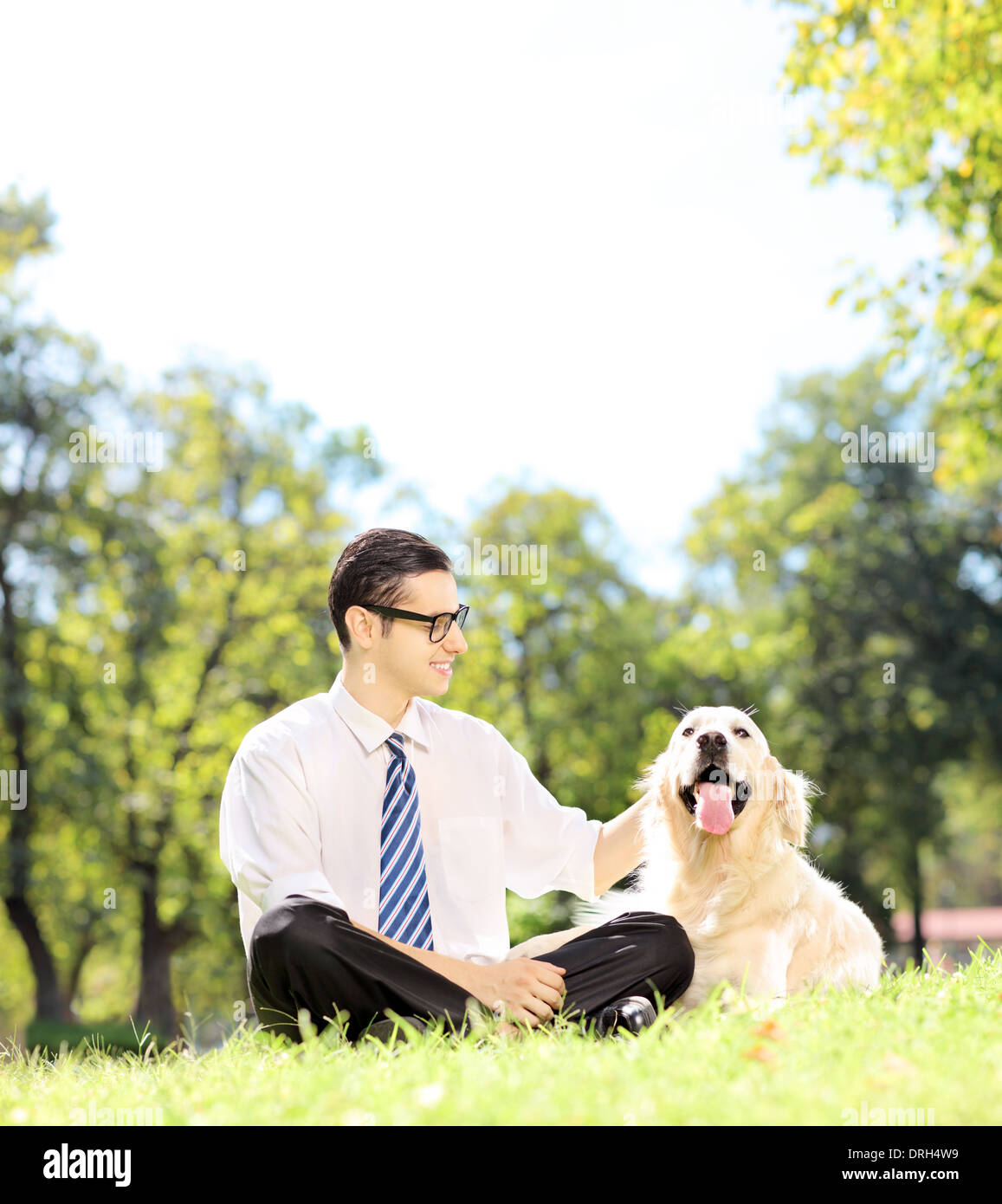 Sorridente giovane imprenditore con il suo cane seduto sul prato in un parco Foto Stock