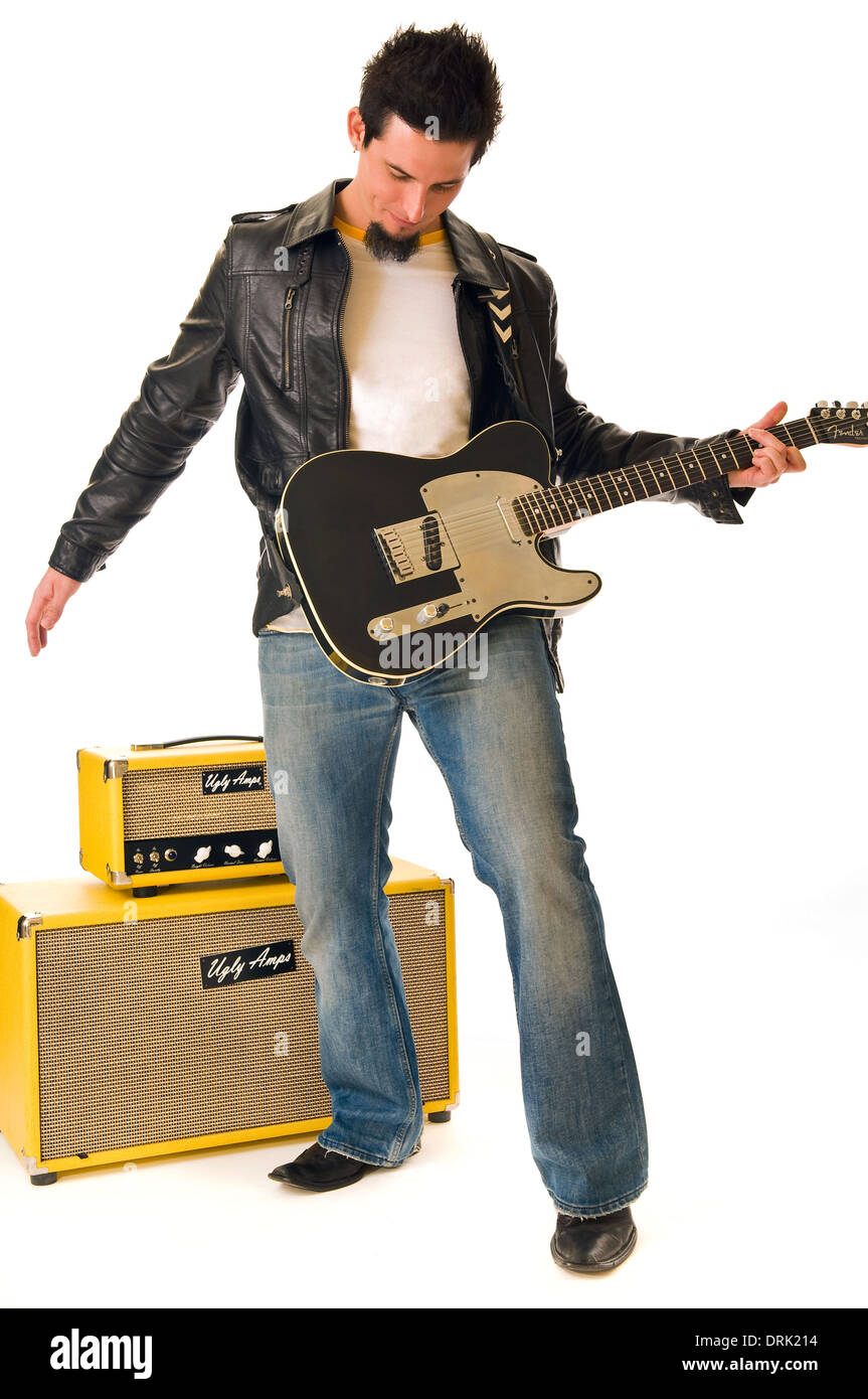 Un musicista rock suonare la chitarra elettrica, indossa una giacca di pelle e jeans blu. Dietro sono impilati giallo amplificatori. Foto Stock