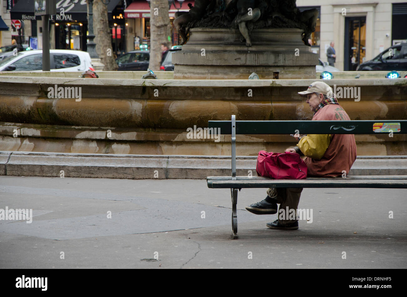 I vecchi donna senzatetto di fumare una sigaretta di fronte a una fontana a Parigi, Francia. Foto Stock