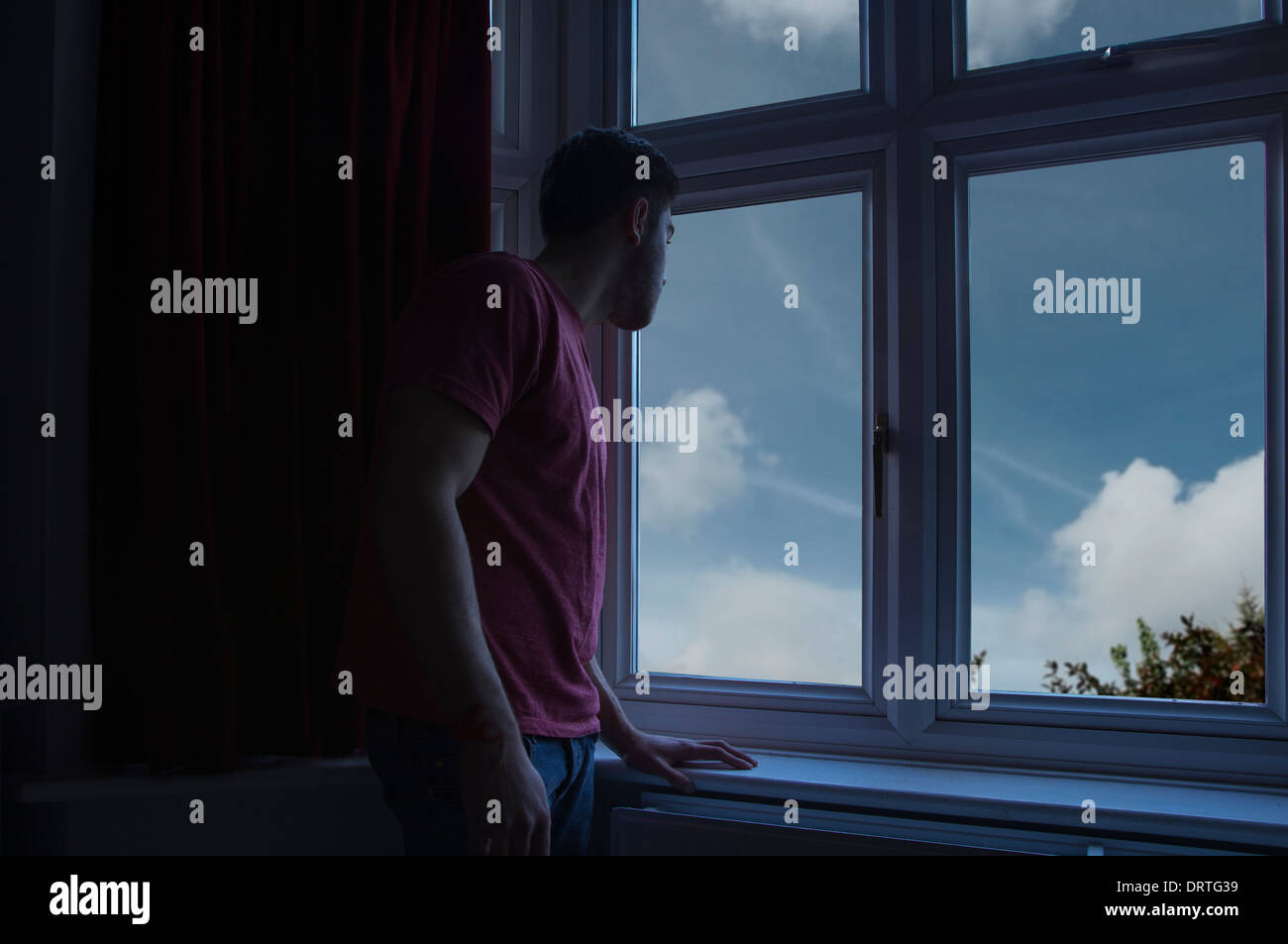 Giovane uomo in una stanza buia guardando fuori attraverso una finestra. Foto Stock