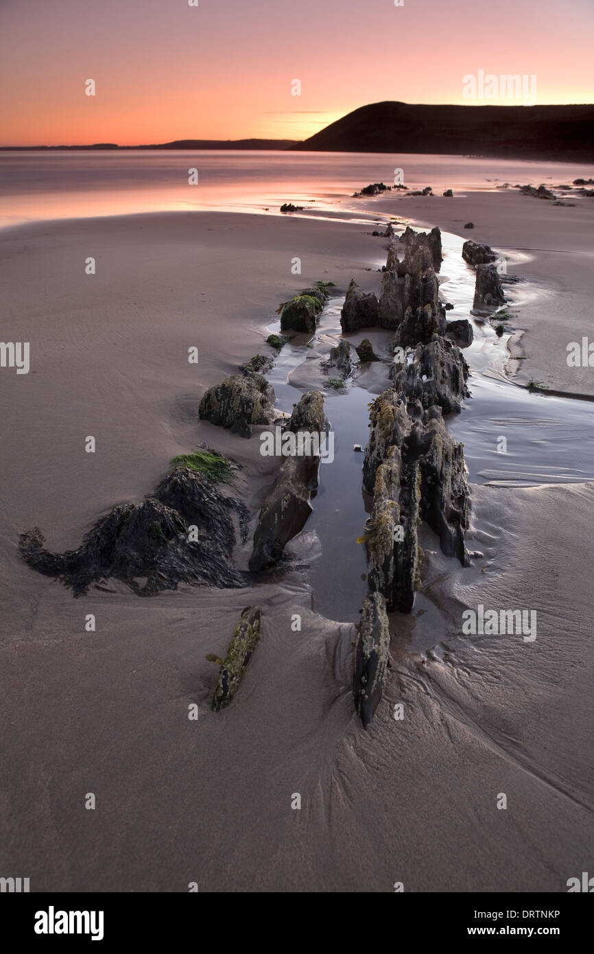Rocce frastagliate che sporgono dalla sabbia a bassa marea durante il tramonto su Manorbier Beach, Pembrokeshire, South Wales, Regno Unito Foto Stock