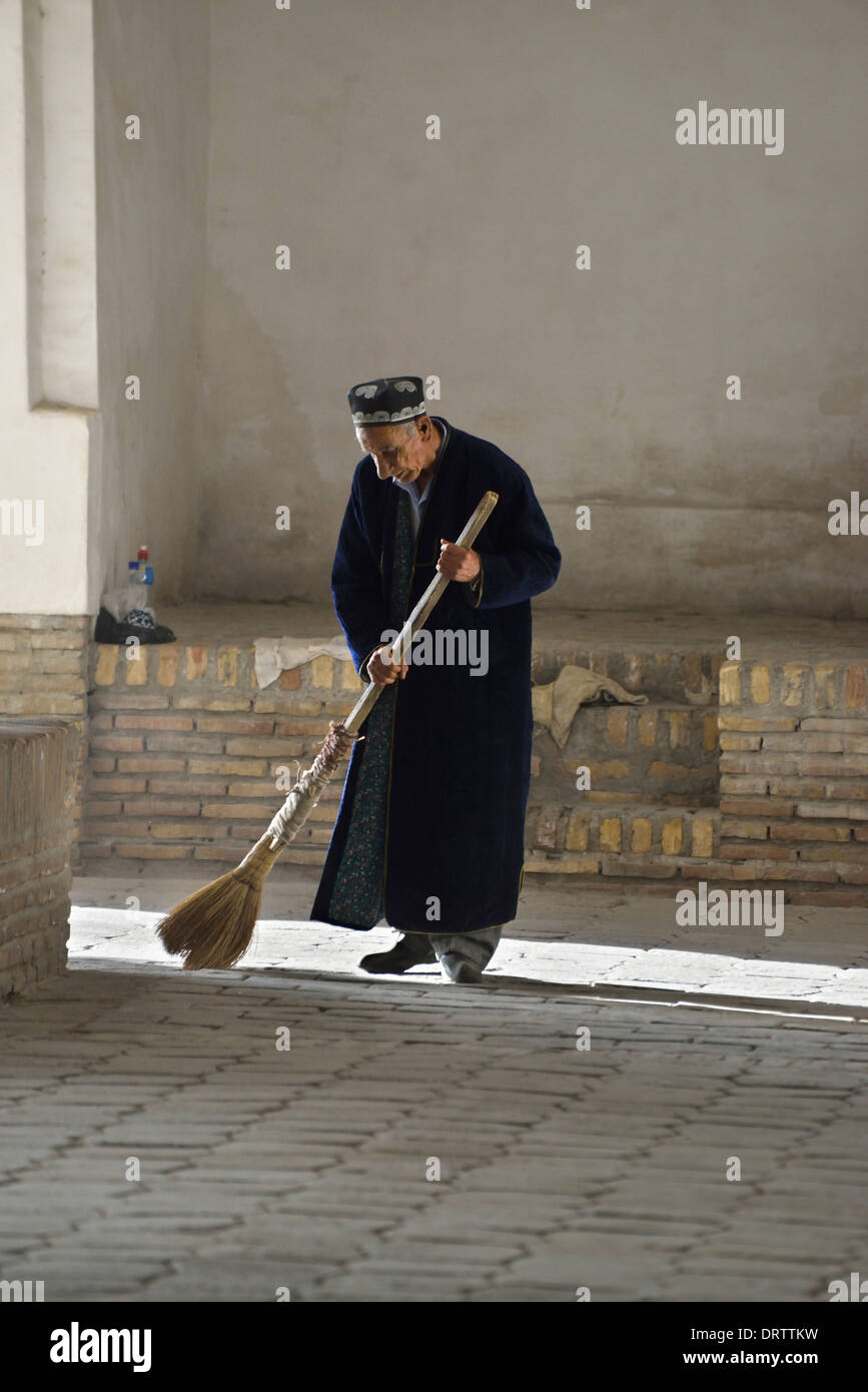Uomo in abito tradizionale spazzare il pavimento bazaar, Bukhara, Uzbekistan Foto Stock