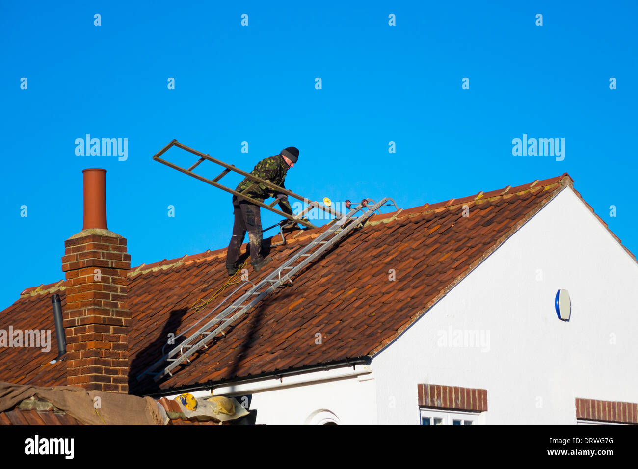 Un uomo su un tetto di casa impostazione delle scale di accesso per la riparazione di vento danneggiato tegole del tetto Foto Stock