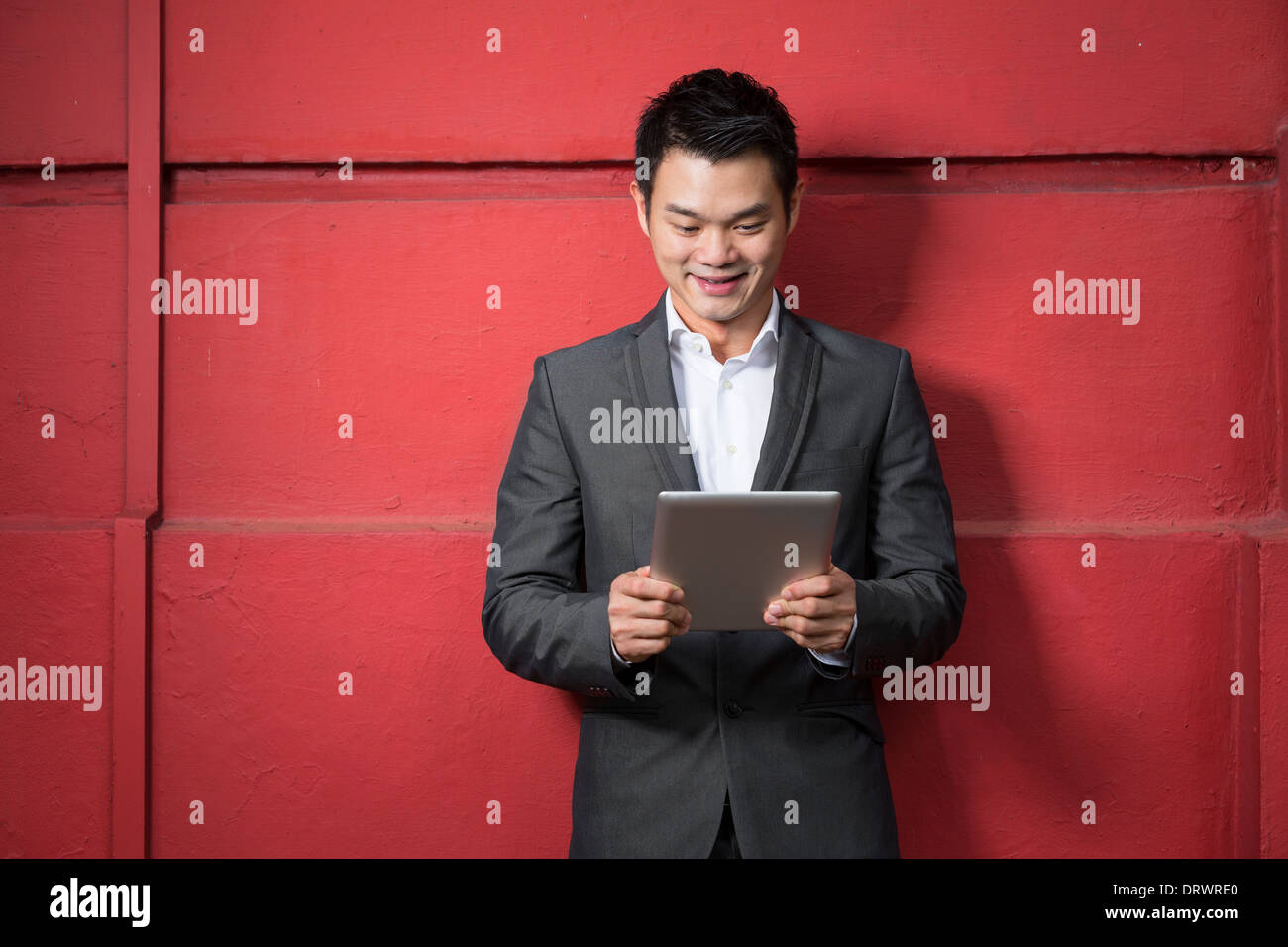 Uomo cinese con un computer tablet. Asian business man la lettura di una tavoletta digitale computer, appoggiata contro una parete rossa Foto Stock