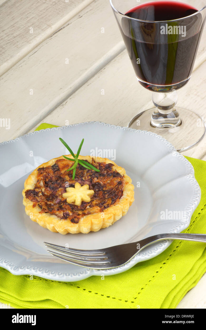 Mini quiche con radicchio trevisano su una lastra grigia e un bicchiere di vino rosso su bianco sullo sfondo di legno Foto Stock