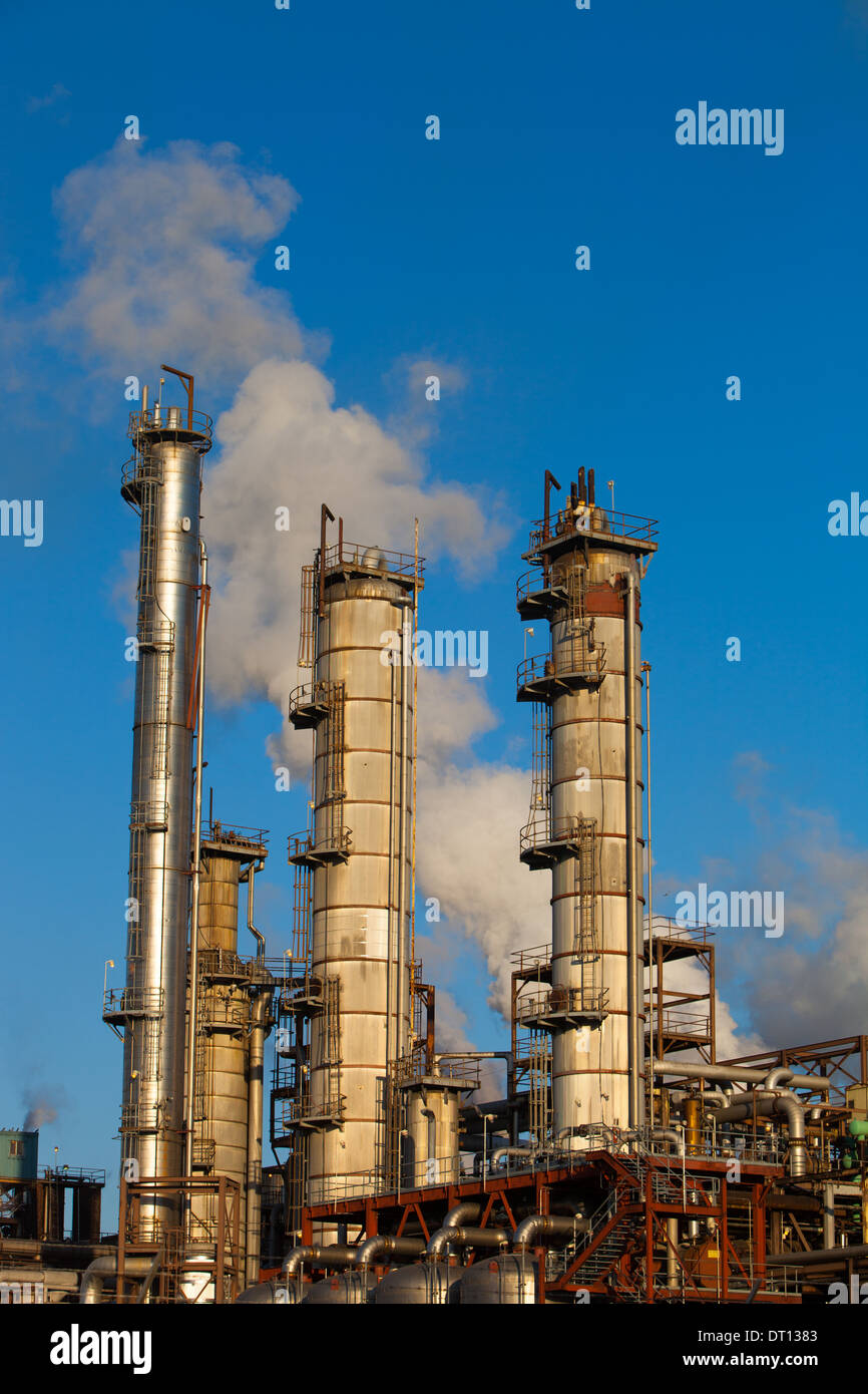 Il vapore sale dalle torri di distillazione e dai camini di una grande raffineria di petrolio sotto un cielo blu. Foto Stock
