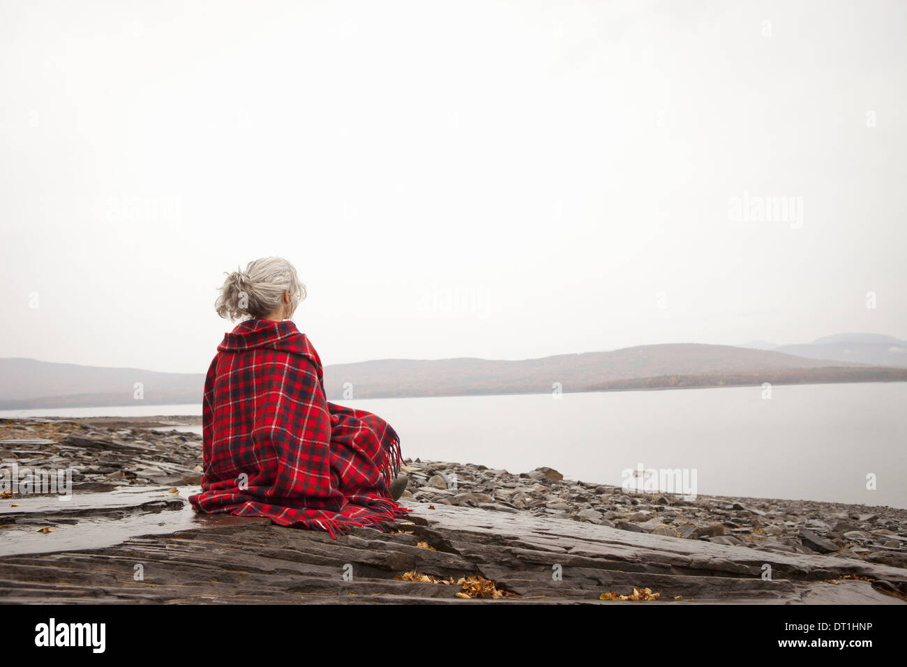 Una donna che guarda sull'acqua sulle rive di un lago calmo avvolto in un tartan rug Foto Stock